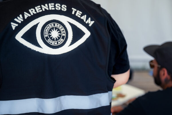 Bild aus dem Festivalgeschehen. Man sieht einen Rücken eines Mitgliedes des Awareness Teams mit der Awareness Team Weste und dem SUMMER BREEZE Awareness Symbol, dem Auge